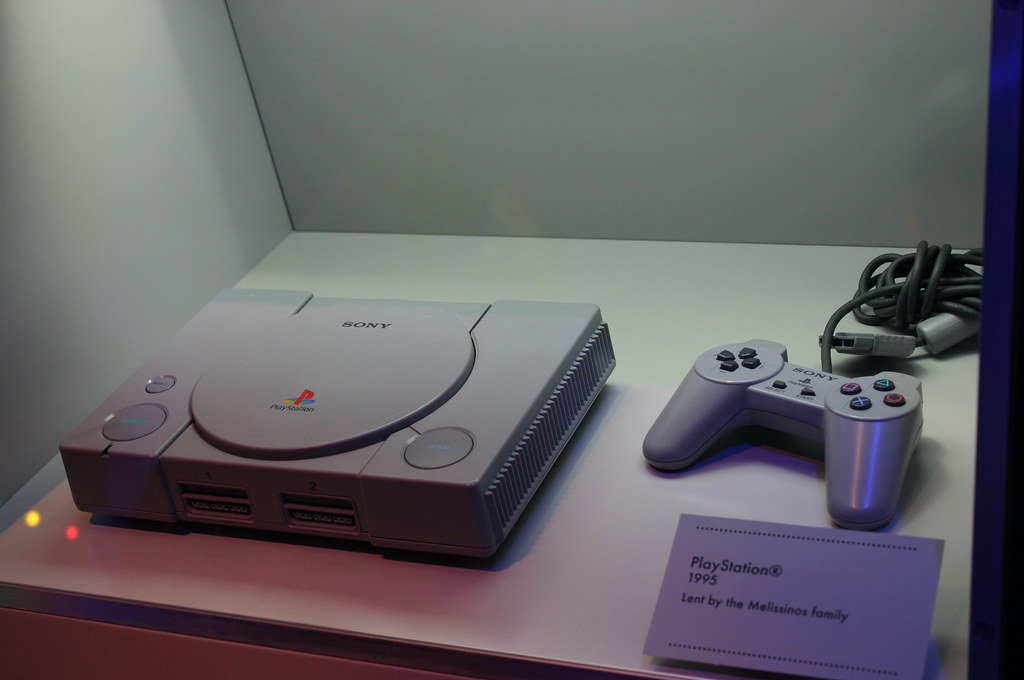 The original PlayStation 1 console and controller on a display stand with a nametag